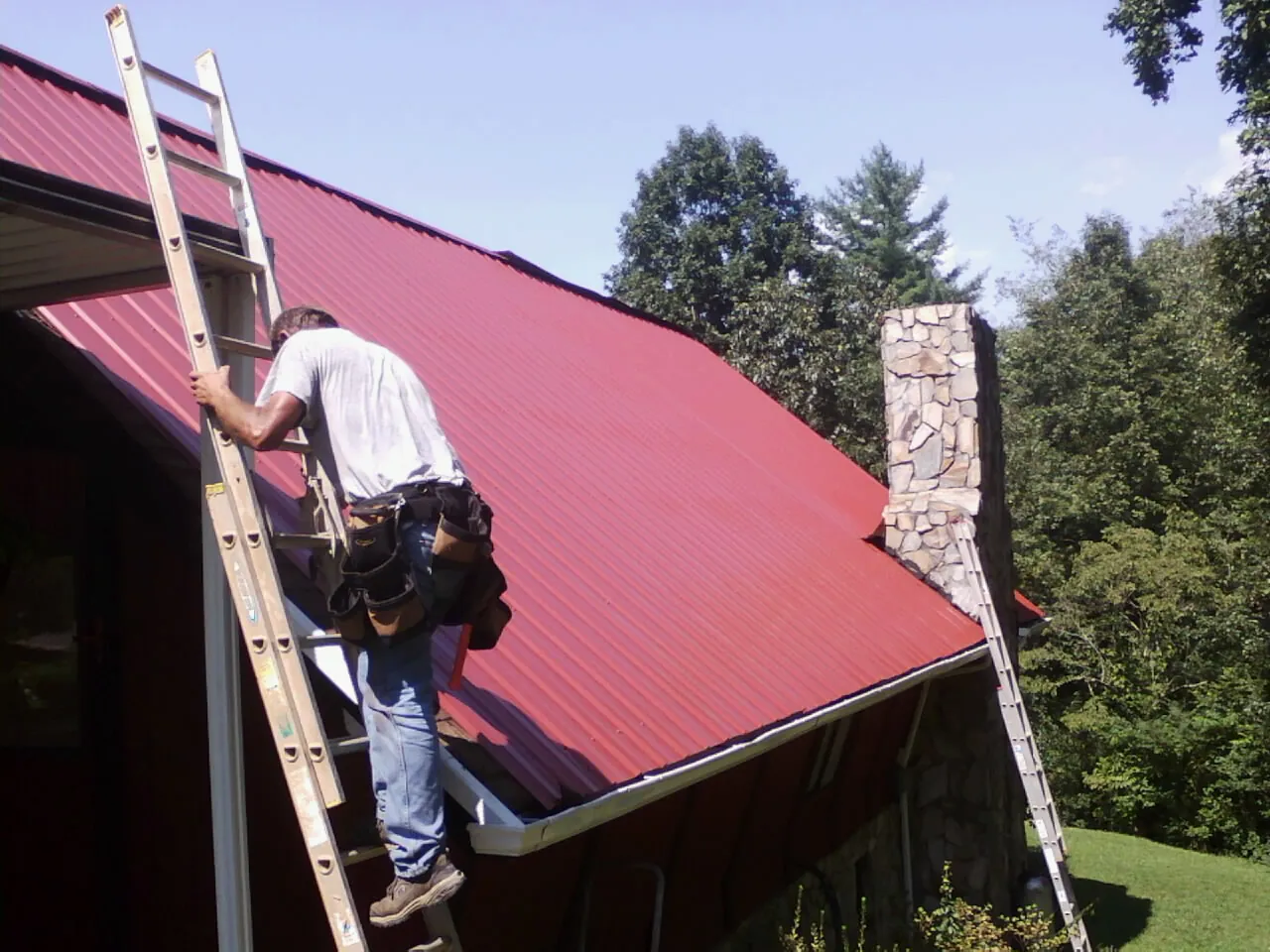 Roofer on a ladder beside a red metal roof and stone chimney — Soffit & Fascia Repair by Roofing Brothers in Huntington